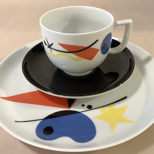 Könnte beinhalten: Ein weißes Keramik-Tassen- und Untertassen-Set mit passendem Teller, das ein abstraktes grafisches Design in Schwarz, Blau, Orange und Gelb aufweist. Die Tasse hat einen quadratischen Henkel, die Untertasse ist einfarbig schwarz und der Teller ist weiß mit bunten Formen.