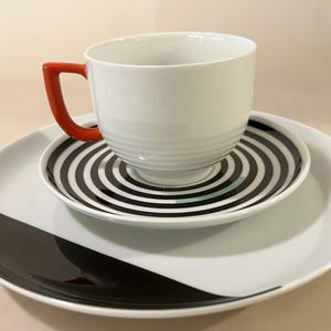 Könnte beinhalten: Eine weiße Keramiktasse mit einem leuchtend orangefarbenen Henkel steht auf einer passenden Untertasse mit konzentrischen schwarzen und weißen Kreisen. Die Untertasse liegt auf einem großen weißen Teller mit einer schwarzen Kurve.