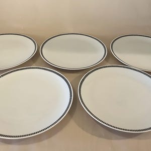 Pode incluir: Cinco travessas ovais brancas com uma borda azul escura em forma de corda. Os pratos são dispostos sobre uma superfície bege clara, mostrando um design limpo e minimalista, adequado para servir alimentos.