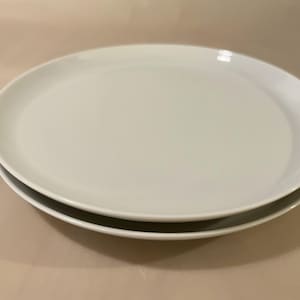 Könnte beinhalten: Ein Stapel aus zwei runden, weißen Keramiktellern mit glatter, glänzender Oberfläche und einem schlichten, klaren Design. Die Teller haben einen leicht erhöhten Rand und eine flache Mitte, was eine einheitliche, durchgehend weiße Farbe ergibt.