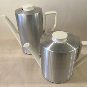 Könnte beinhalten: Zwei silberfarbene Metall-Teekannen mit weißen Ausgüssen, Griffen und Deckeln. Die Teekannen haben einen zylindrischen Körper und ein modernes Design. Eine Teekanne ist etwas größer als die andere.