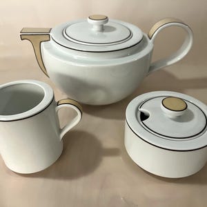 Op de afbeelding: Een wit porseleinen theeservies met een crèmekleurige rand. De set bevat een theepot, een roomkan en een suikerpot met deksel.