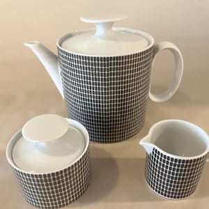 Pode incluir: Um conjunto de chá com padrão a preto e branco. O conjunto inclui uma chaleira com tampa, um açucareiro com tampa e um cremeira. A chaleira tem um bico e uma pega curvos. O açucareiro e a cremeira têm um design simples e arredondado.