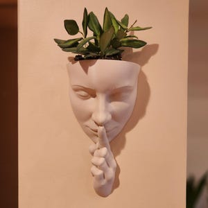 May include: A white wall planter shaped like a human face, with a finger pressed to the lips in a 'shush' gesture. The planter holds green leafy plants. The face has closed eyes and is mounted on a light-colored wall.
