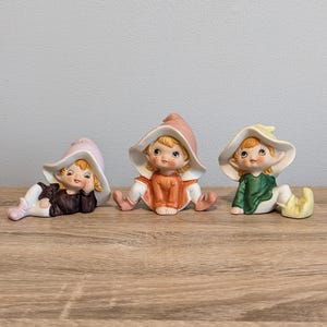 May include: Three ceramic figurines of seated elves with large, floppy hats. One elf wears a pink hat and brown outfit, another has an orange outfit and pink hat, and the third wears a green top and yellow hat.