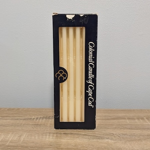 May include: A box of six Colonial Candle of Cape Cod candles. The candles are white and have a gold shimmer. The box is black with gold lettering.