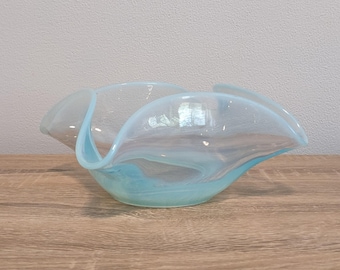 May include: A decorative glass bowl with a wavy, organic shape. The bowl features a blend of light blue and clear glass, creating a marbled effect. The bowl is sitting on a wooden surface.