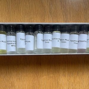 May include: A white box containing twelve small glass bottles with black caps and white labels. The labels feature text such as "Egyptian Musk" and "Gold". The bottles are filled with various colored liquids.