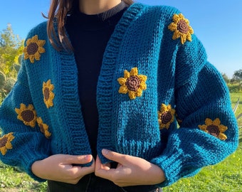 May include: Teal blue, chunky knit cardigan adorned with yellow and brown sunflower appliqués. The cropped sweater has a V-neck and long, puffy sleeves. The cardigan is being held closed by the wearer.