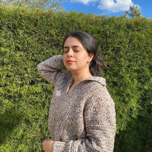 May include: A woman wearing a beige, brown, and white crocheted sweater with a v-neck and long sleeves. She is standing in front of a green hedge.