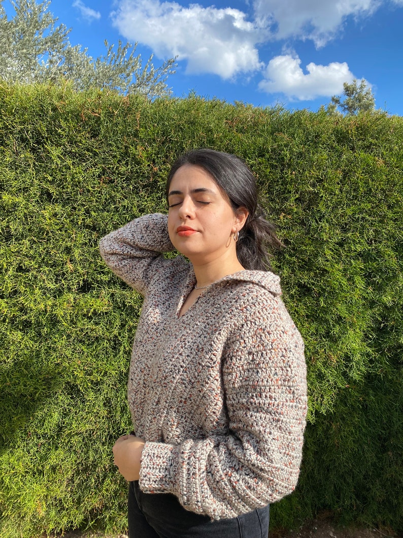 May include: A woman wearing a beige, brown, and white crocheted sweater with a v-neck and long sleeves. She is standing in front of a green hedge.