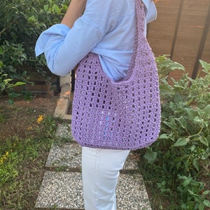 May include: A lavender crocheted tote bag with a long strap. The bag is being worn over the shoulder of a person wearing a light blue shirt and white pants.