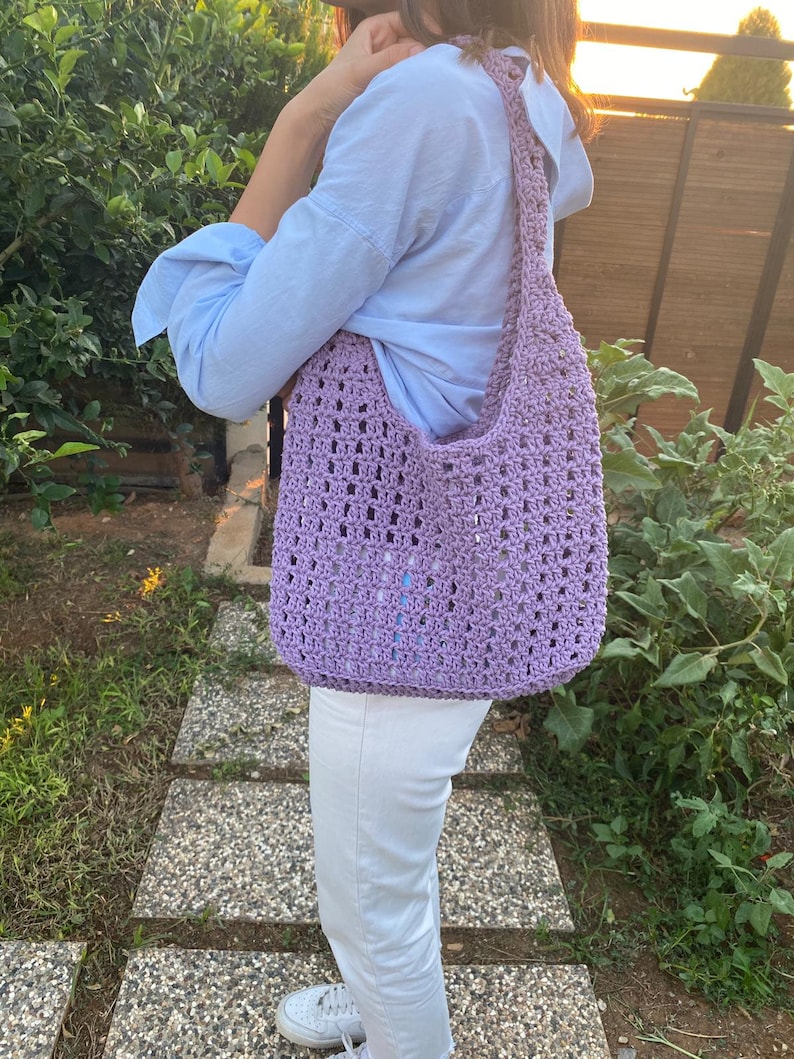 May include: A lavender crocheted tote bag with a long strap. The bag is being worn over the shoulder of a person wearing a light blue shirt and white pants.