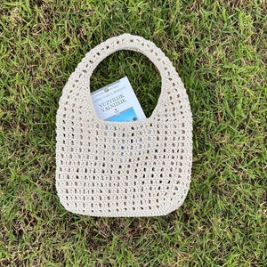 May include: A beige crochet tote bag with a book titled "Y&Uuml;ZYILLIK YALNIZLIK" inside. The bag is sitting on a bed of green grass.