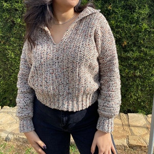 May include: A beige, brown, and red speckled knit sweater with a collared neckline. The sweater has a cropped fit and long sleeves.