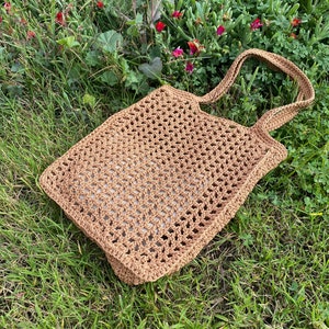 May include: A brown crocheted tote bag with a square shape and two long straps. The bag is lying on a bed of green grass.