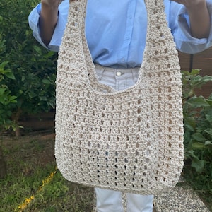 May include: A beige crocheted tote bag with a large open top. The bag is made with a simple stitch pattern and has a relaxed, casual look.