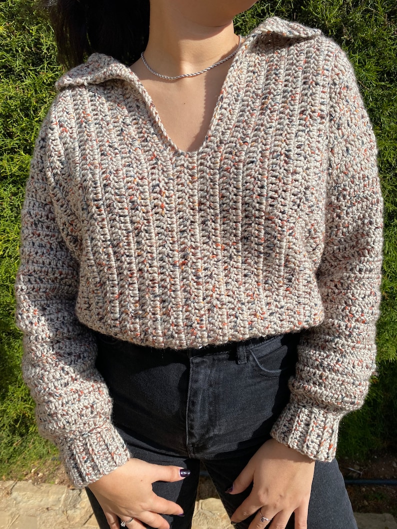 May include: A beige crocheted sweater with a v-neck and long sleeves. The sweater has a speckled pattern with brown, black, and white yarn.