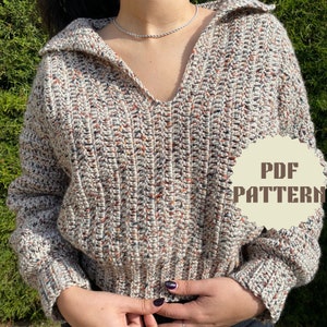 May include: A crocheted sweater with a v-neck and long sleeves. The sweater is made with a speckled yarn in shades of white, brown, and orange. The sweater has a relaxed fit and a slightly cropped length. The image includes the text "PDF PATTERN".