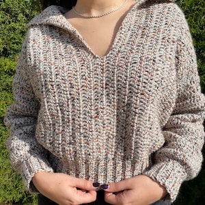 May include: A beige, brown, and white speckled crochet sweater with a v-neck and a collar. The sweater has long sleeves and a ribbed hem.