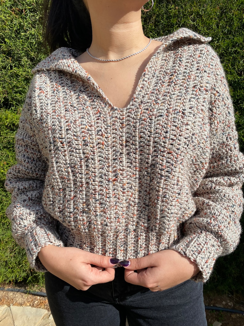 May include: A beige, brown, and white speckled crochet sweater with a v-neck and a collar. The sweater has long sleeves and a ribbed hem.