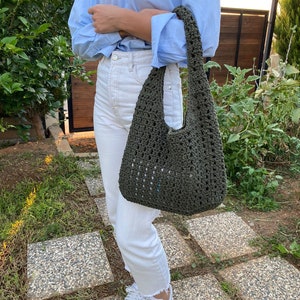 May include: A green crocheted tote bag with a wide, open top and a single strap. The bag is being held by a person wearing white jeans and a light blue shirt.