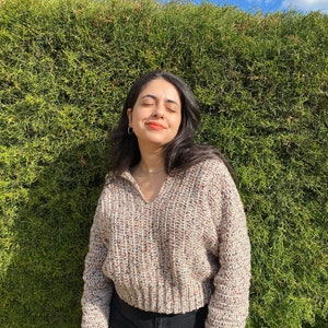 May include: A woman wearing a beige, brown, and white speckled cropped sweater with long sleeves and a v-neck. She is wearing black jeans and has her eyes closed.