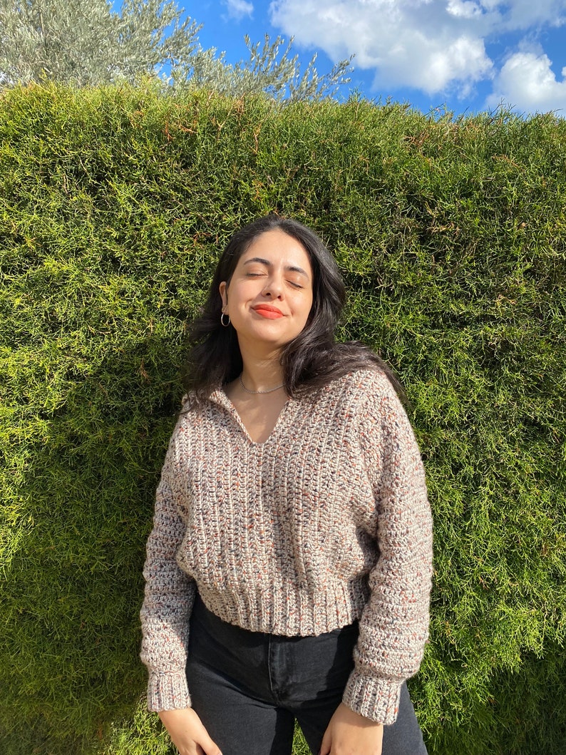 May include: A woman wearing a beige, brown, and white speckled cropped sweater with long sleeves and a v-neck. She is wearing black jeans and has her eyes closed.