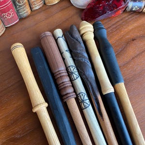 May include: A collection of handcrafted wooden wands in various shades of brown and black. Each wand features unique designs, including carved textures and painted details. The wands are arranged on a wooden surface, showcasing their individual craftsmanship.