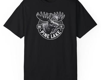 以下が含まれることがあります： 黒いTシャツ。大きな角を持つヘラジカの頭部の白黒イラストが描かれています。「PINE LAKE」と「OMA WI」の文字が円形フレームに収められています。
