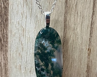 May include: A silver chain necklace with an oval pendant. The pendant is a polished stone with a green and white moss-like pattern. The necklace is displayed against a wooden background.
