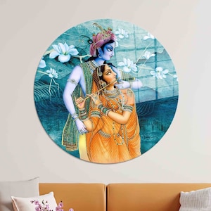 May include: A circular wall art piece featuring a depiction of Krishna and Radha, a Hindu deity couple, in a romantic embrace. The artwork is painted in a traditional Indian style with vibrant colors and intricate details. The background features a blue and green watercolor wash with white lotus flowers.