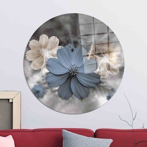 May include: A round wall art piece featuring a blue flower with white accents and two beige flowers with brown centers. The flowers are set against a blurred background of gray and white.