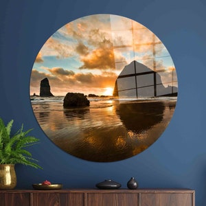 May include: A circular print featuring a scenic sunset over a beach with a large rock formation in the distance. The sky is a vibrant orange and pink, with clouds reflecting in the water.