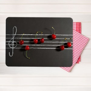 Pode incluir: Um quadro-negro preto com linhas de pauta brancas e uma clave de sol. Cerejas vermelhas brilhantes são dispostas nas linhas para se assemelharem a notas musicais. Uma toalha de mesa xadrez vermelha e branca está no canto.