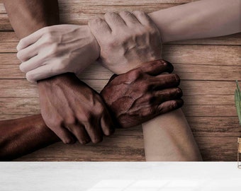 May include: Four hands of different skin tones are clasped together in a circle, symbolizing unity and diversity. The hands are against a wooden background.