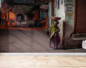May include: A woman in a green shawl and purple dress carries a basket on her head as she walks down a narrow street in India. The street is lined with buildings, and there is a sign for "Hotel Paramount Palace" on one of the buildings.