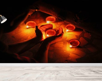 May include: A person's hands cupped around a small, lit candle, with several other lit candles scattered on the floor. The scene is dimly lit, with a single lamp in the background.