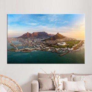 Könnte beinhalten: Eine Luftaufnahme von Kapstadt, Südafrika, mit dem Tafelberg und dem Löwenkopf im Hintergrund. Die Stadt erstreckt sich darunter, mit dem Meer im Vordergrund. Ein großes Stadion ist in der Mitte des Bildes zu sehen.