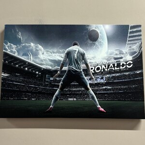 Könnte beinhalten: Leinwanddruck mit einem Fußballspieler in einem Stadion. Der Spieler, der ein hellblaues Trikot mit der Nummer 7 trägt, ist von hinten zu sehen. Der Druck enthält den Text "Cristiano Ronaldo" und "The Best in the World". Der Hintergrund zeigt ein Stadion voller Zuschauer.