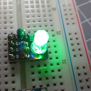 以下が含まれることがあります： ブレッドボードに緑色のLEDライトが点灯しています。ライトは、「G1」、「G」、「B」、「R」の文字が印刷された小さな回路基板に接続されています。回路基板には「SUMU RGB BREAD BOARD」とラベル付けされています。