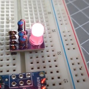 Könnte beinhalten: Eine rote LED-Leuchte leuchtet auf einem Steckbrett mit einer kleinen Platine, die daran befestigt ist. Die Platine hat ein paar blaue Widerstände und einen kleinen Chip.