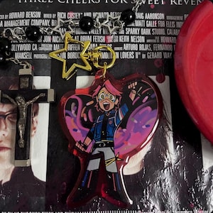 以下が含まれることがあります： 黒と銀のロザリオで、黒いビーズと銀のチャームが付いています。チャームには星と十字架があります。ロザリオは、赤とピンクのハート型のチャームにぶら下がっています。チャームには、青と白のジャケットと黄色のズボンを着た漫画のキャラクターが描かれています。キャラクターはマイクを持っていて、ピンクと紫の背景があります。