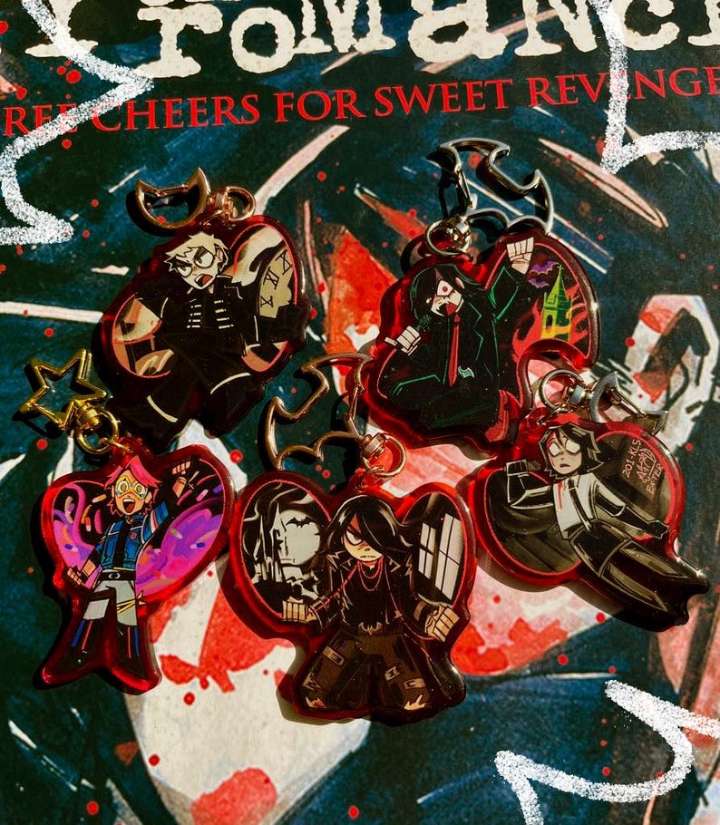 以下が含まれることがあります： アニメのキャラクターが描かれた赤いアクリルキーホルダーが5つ。それぞれのキーホルダーには、黒髪で黒服を着た異なるキャラクターが描かれています。キャラクターはさまざまなポーズをとっています。キーホルダーは、赤と白の文字が書かれた暗い背景の上に置かれています。