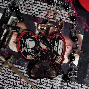 以下が含まれることがあります： 赤色のハート型のキーホルダー。黒と白の漫画のイラストが描かれており、2人の人物が描かれている。左の人物は黒いジャケットを着ており、右の人物は黒いジャケットと赤いネックレスを着ている。