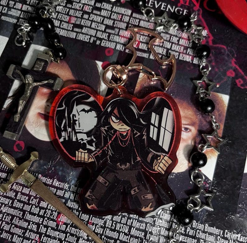 以下が含まれることがあります： 赤色のハート型のキーホルダー。黒と白の漫画のイラストが描かれており、2人の人物が描かれている。左の人物は黒いジャケットを着ており、右の人物は黒いジャケットと赤いネックレスを着ている。