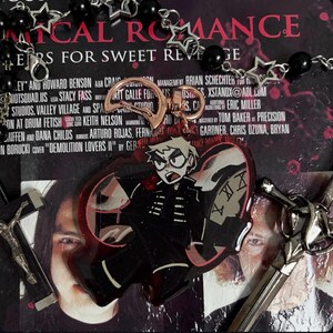 以下が含まれることがあります： 黒い羽根と赤い羽根を持つ蝶の形をしたチャーム。黒い服を着た漫画のキャラクターと時計の文字盤が羽根に描かれている。チャームは、「MYTHICAL ROMANCE」と「ERS FOR SWEET REVENGE」と書かれたCDの上に置かれている。