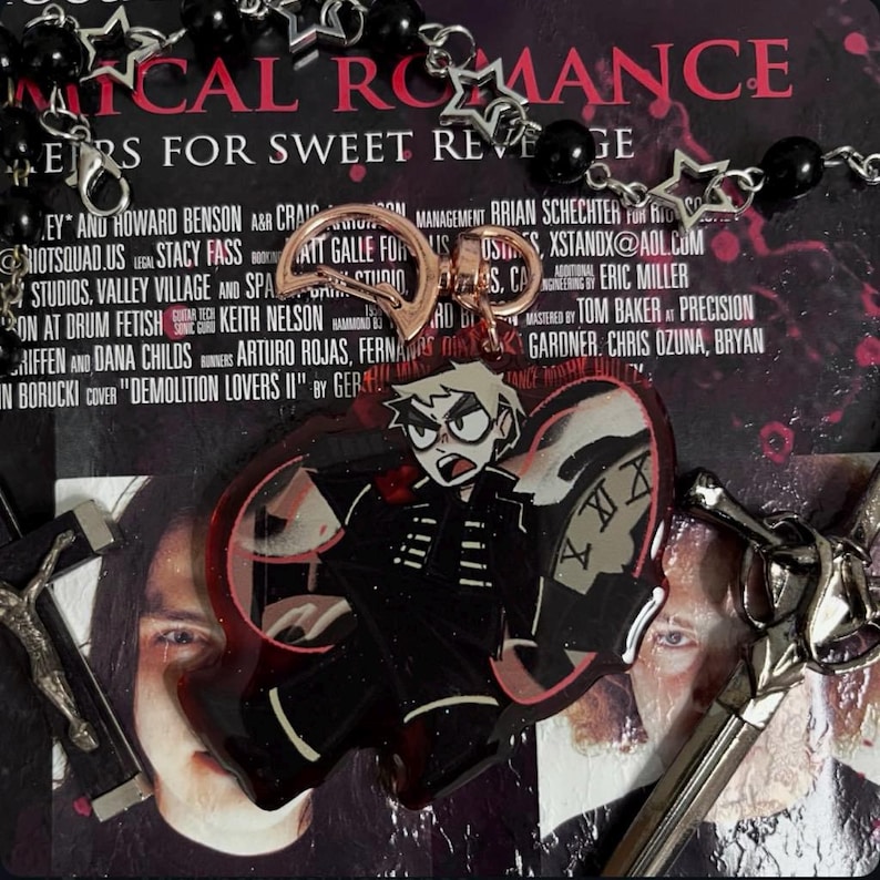 以下が含まれることがあります： 黒い羽根と赤い羽根を持つ蝶の形をしたチャーム。黒い服を着た漫画のキャラクターと時計の文字盤が羽根に描かれている。チャームは、「MYTHICAL ROMANCE」と「ERS FOR SWEET REVENGE」と書かれたCDの上に置かれている。