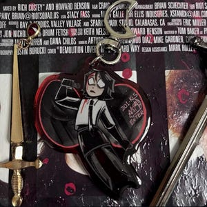 以下が含まれることがあります： 赤い目をした白黒の漫画のキャラクターが、赤いシミのついた白いシャツを着ています。キャラクターはナイフを持っており、赤いハートの前に立っています。キャラクターは、銀色の金属製のリングが付いたキーホルダーに付いています。キーホルダーは、黒と赤の背景に「201-KL5 EXTER」という文字が書かれています。