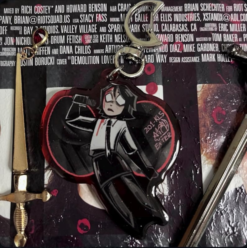 以下が含まれることがあります： 赤い目をした白黒の漫画のキャラクターが、赤いシミのついた白いシャツを着ています。キャラクターはナイフを持っており、赤いハートの前に立っています。キャラクターは、銀色の金属製のリングが付いたキーホルダーに付いています。キーホルダーは、黒と赤の背景に「201-KL5 EXTER」という文字が書かれています。